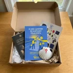 O Kit O Meu Caminho Pela Costa ainda dentro da caixa, com o Guia do Caminho da Costa, o postal com a credencial, o postal da Checklist e a vieira do peregrino.